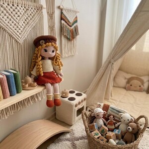 May include: A cozy children's room with a handmade crochet doll wearing a brown hat and red skirt. A woven basket holds stuffed animals and wooden blocks. A wooden balance board and a small play kitchen add to the playful atmosphere.