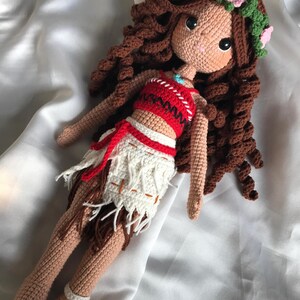 May include: A handmade crochet doll with brown curly hair, wearing a red and white top, and a white skirt with fringe. The doll has a floral crown and is lying on a white satin surface. The doll's skin is tan, and it has large black eyes.
