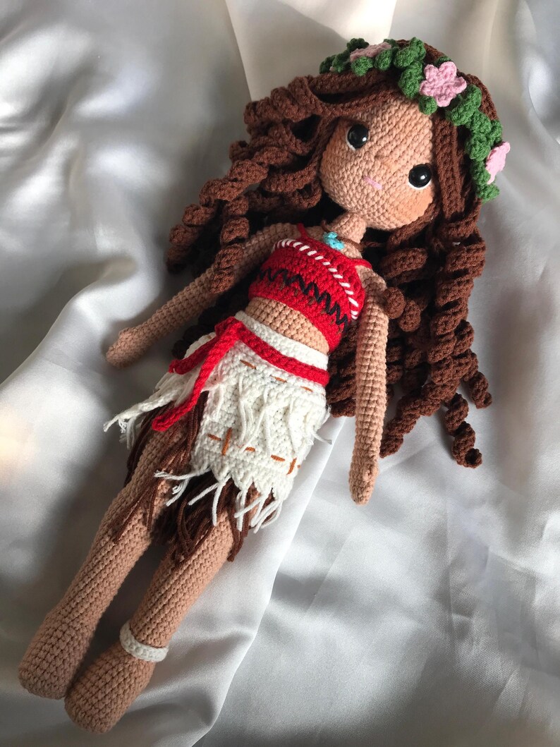 May include: A handmade crochet doll with brown curly hair, wearing a red and white top, and a white skirt with fringe. The doll has a floral crown and is lying on a white satin surface. The doll's skin is tan, and it has large black eyes.
