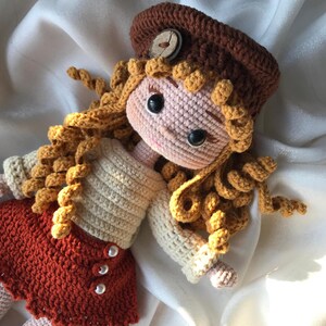 May include: A handmade crochet doll with a brown hat and curly blonde hair. The doll wears a cream-colored sweater and a rust-colored skirt with pearl buttons. The doll's face is pink with black eyes.