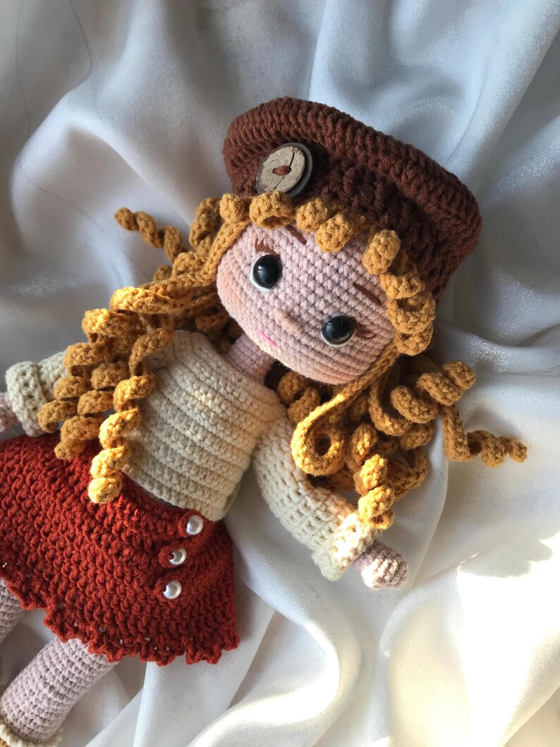 May include: A handmade crochet doll with a brown hat and curly blonde hair. The doll wears a cream-colored sweater and a rust-colored skirt with pearl buttons. The doll's face is pink with black eyes.