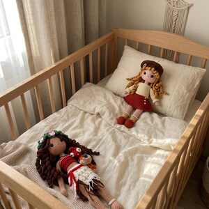May include: Two handmade crochet dolls rest on a crib. One doll has long brown hair, a floral crown, and a red and white outfit. The other doll has blonde hair, a brown beret, and a red skirt. The crib is made of light-colored wood.