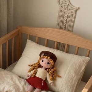 May include: A handmade crochet doll with long, curly blonde hair, wearing a brown beret, cream-colored sweater, and a red skirt. The doll is resting on a white pillow in a wooden crib. The doll has rosy cheeks and is wearing red shoes.
