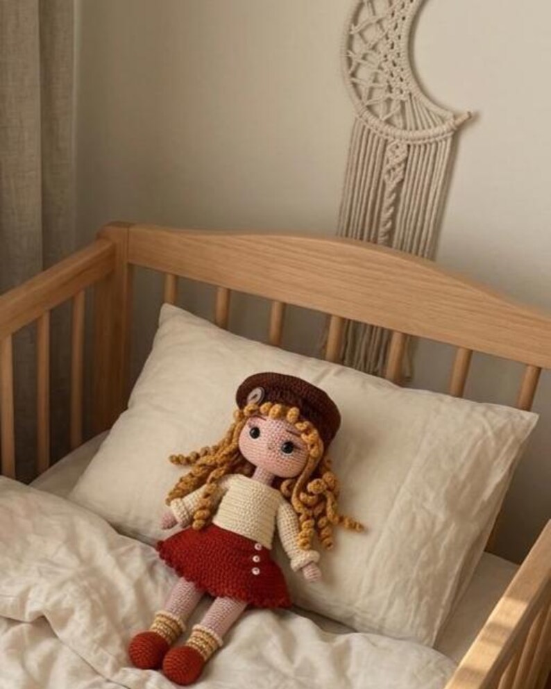 May include: A handmade crochet doll with long, curly blonde hair, wearing a brown beret, cream-colored sweater, and a red skirt. The doll is resting on a white pillow in a wooden crib. The doll has rosy cheeks and is wearing red shoes.