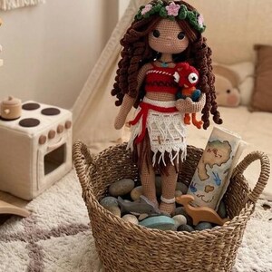 May include: A handmade crochet doll with brown curly hair, wearing a red and white outfit, and holding a red plush toy. The doll is in a woven basket filled with stones and wooden toys. A map is also in the basket.