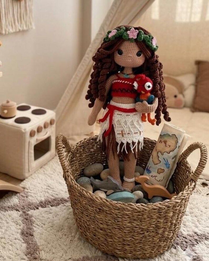 May include: A handmade crochet doll with brown curly hair, wearing a red and white outfit, and holding a red plush toy. The doll is in a woven basket filled with stones and wooden toys. A map is also in the basket.