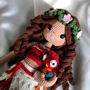 May include: A handmade crochet doll with brown curly hair, a floral crown, and a tan face. The doll wears a red and white outfit and holds a small, colorful crochet bird. The doll's eyes are large and black.