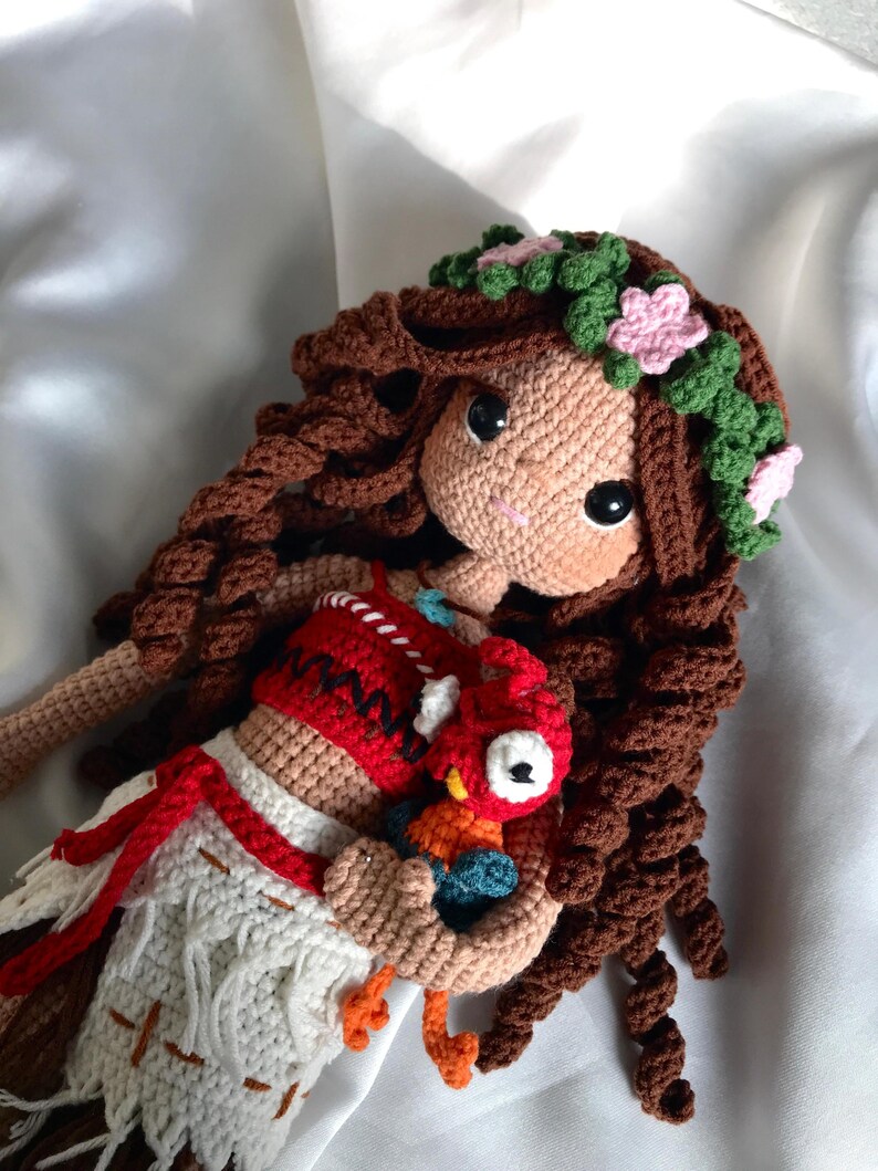 May include: A handmade crochet doll with brown curly hair, a floral crown, and a tan face. The doll wears a red and white outfit and holds a small, colorful crochet bird. The doll's eyes are large and black.