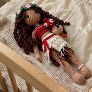 May include: A handmade crochet doll with brown hair, a floral headband, and a red and white outfit. The doll is holding a small red and orange bird. The doll is lying on a beige blanket.