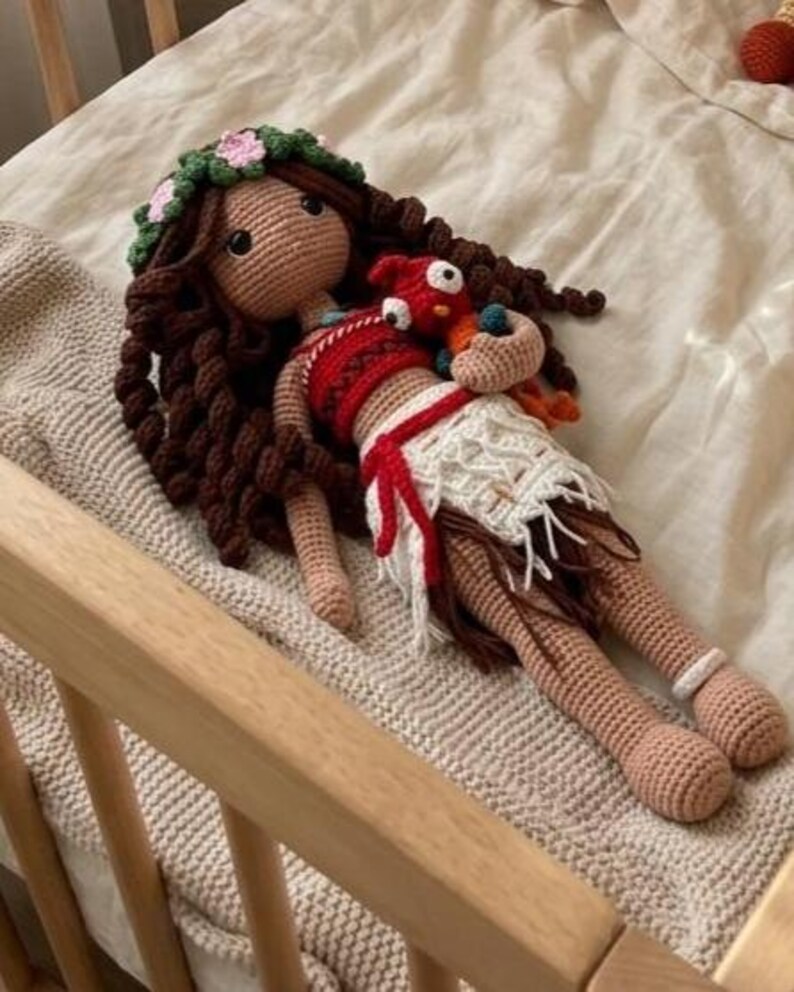 May include: A handmade crochet doll with brown hair, a floral headband, and a red and white outfit. The doll is holding a small red and orange bird. The doll is lying on a beige blanket.