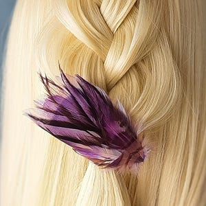 May include: A close-up shot of blonde hair styled in a braid, with a purple feather hair accessory. The feather features shades of dark purple and brown, contrasting with the light hair. The background is a plain, muted blue.