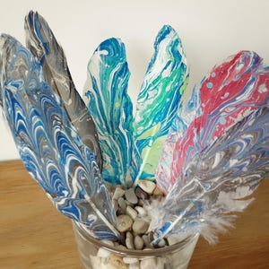 May include: A collection of feathers featuring marbled patterns in blue, grey, white, green, and red. The feathers are arranged upright in a clear glass container filled with small, smooth, light-coloured pebbles, creating a display of vibrant colours.