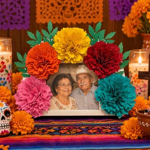 May include: A white rectangular picture frame containing a photograph of an older couple, decorated with large, vibrant paper flowers in yellow, orange, red, pink, and teal, accented with green paper leaves attached to the top and sides.