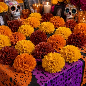 May include: A festive Day of the Dead altar display featuring numerous bright orange and deep red marigold flowers arranged on a black table covered with orange and purple paper cutouts, two decorated sugar skulls, and several lit candles.