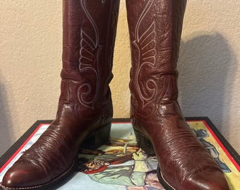 May include: A pair of burgundy leather cowboy boots with decorative white stitching. The boots have a pointed toe and a stacked heel. The leather has a textured appearance. The boots are displayed on a box with a colorful illustration.
