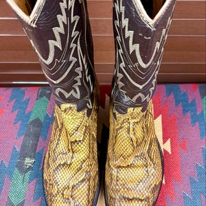 May include: A pair of tall cowboy boots featuring dark brown leather shafts with white decorative stitching and pointed toes covered in a textured yellow and brown snakeskin pattern. The interior lining is tan leather with visible text.