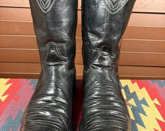 Peut inclure: Une paire de bottes de cow-boy en cuir noir avec un motif texturé en peau de lézard sur l'empeigne et des tiges en cuir lisse. Les bottes présentent des surpiqûres blanches décoratives sur le haut et des tirettes latérales.