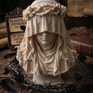 May include: A beige, speckled sculpture of a figure wearing a crown of thorns and a veil that incorporates architectural gothic cathedral elements. The bust features a serene facial expression, with the veil draping over the shoulders and chest.