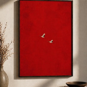 May include: Framed art print depicting two white birds soaring against a bold red backdrop. The artwork, in a black frame, has a minimalist style and a textured finish. The print is displayed on a wooden surface.