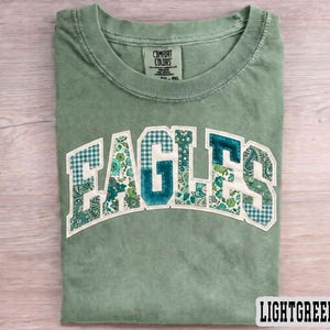 May include: Light green t-shirt featuring the word "EAGLES" in a patchwork design. The letters combine floral, paisley, and gingham patterns in green and teal hues. The shirt has a crew neck and the text "LIGHTGREEN" is at the bottom.