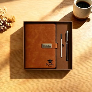May include: A brown leather journal and pen set in a gift box. The journal has a metal clasp and the text "Dr. Joshua" with a graduation cap. A silver pen and a black pen are included. A cup of coffee sits nearby.
