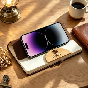 May include: A smartphone sits on a white and brown notebook, with a wooden plaque featuring a graduation cap and the text "Dr. Gavin, Ph.D 2020". A coffee cup and a leather notebook are also on the wooden surface.
