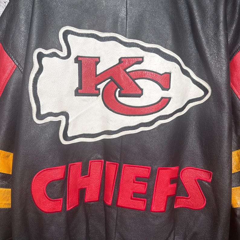 May include: Black leather jacket featuring the Kansas City Chiefs logo. The logo is a white arrowhead with a red "K" and a red "C" inside. The word "CHIEFS" is embroidered in red below the logo. Red and yellow accents are visible.
