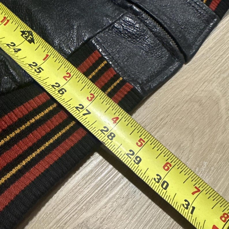 May include: Close-up of a black leather jacket sleeve with a black, red, and gold striped cuff. A yellow measuring tape shows the sleeve's length in inches. The jacket appears to be vintage.
