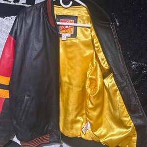 May include: Black leather bomber jacket with red and yellow accents on the sleeves and trim. The interior lining is a bright yellow satin. The jacket is hanging on a white hanger.