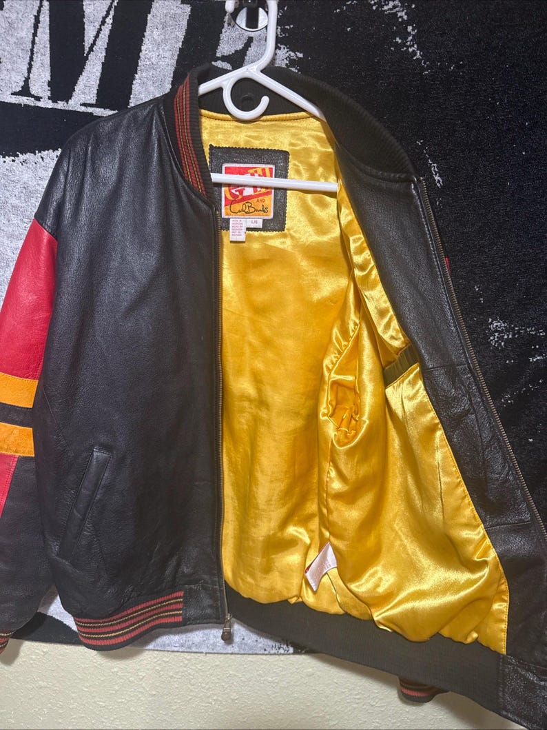 May include: Black leather bomber jacket with red and yellow accents on the sleeves and trim. The interior lining is a bright yellow satin. The jacket is hanging on a white hanger.