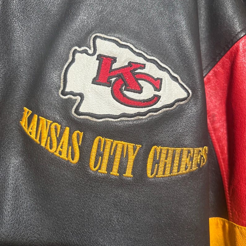 May include: Black and red leather jacket with the Kansas City Chiefs logo patch. The patch features a white arrowhead with a red "KC" logo. The words "KANSAS CITY CHIEFS" are embroidered in gold.