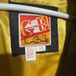 May include: Close-up of a black leather jacket label with the text "GH AND Carl Banks." The label is rectangular with a red and yellow design. The jacket's interior is lined with yellow satin. The label also includes the text "MADE IN INDONESIA."