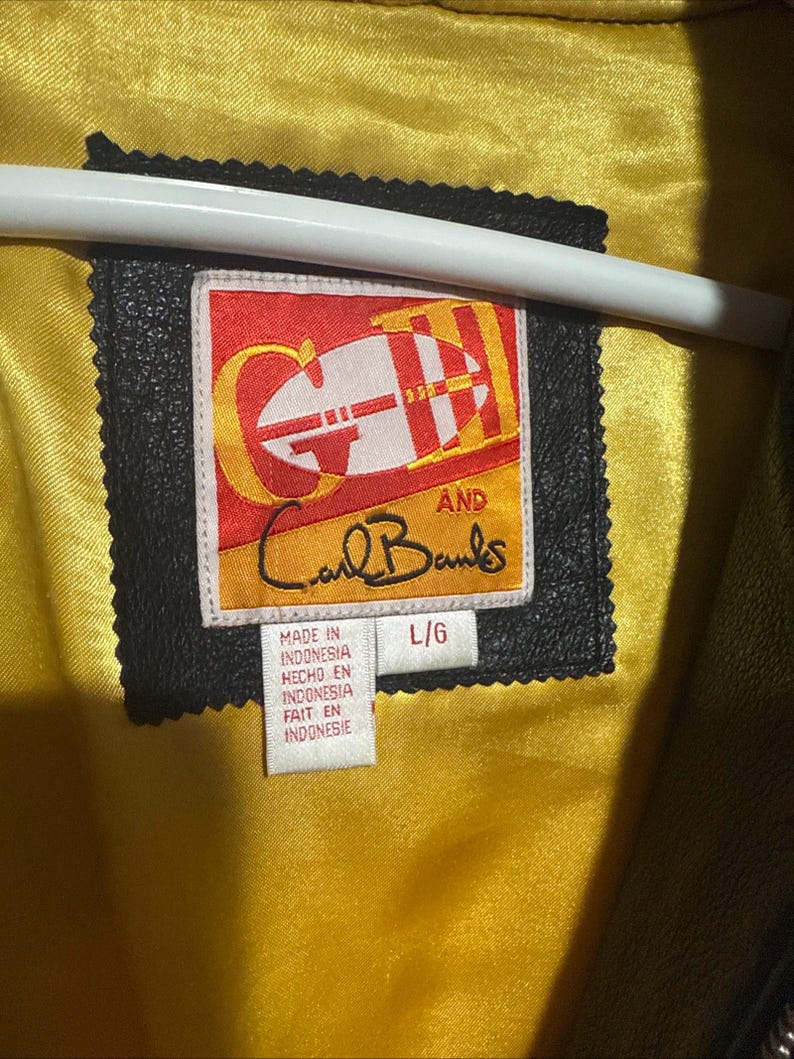 May include: Close-up of a black leather jacket label with the text "GH AND Carl Banks." The label is rectangular with a red and yellow design. The jacket's interior is lined with yellow satin. The label also includes the text "MADE IN INDONESIA."
