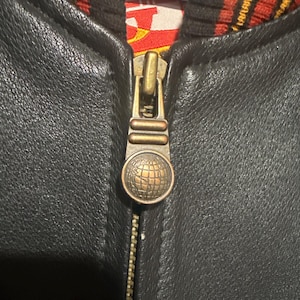 May include: Close-up of a black leather jacket zipper. The zipper is gold-toned with a globe design on the pull. The jacket's interior lining is visible, featuring a red, yellow, and black pattern.