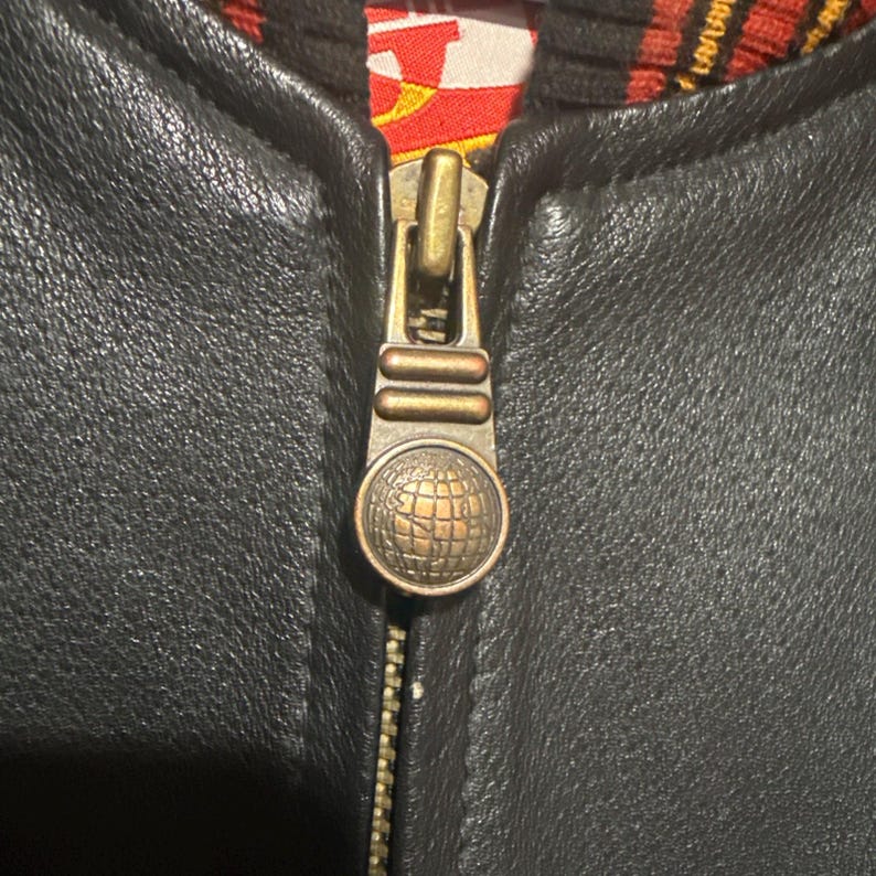 May include: Close-up of a black leather jacket zipper. The zipper is gold-toned with a globe design on the pull. The jacket's interior lining is visible, featuring a red, yellow, and black pattern.
