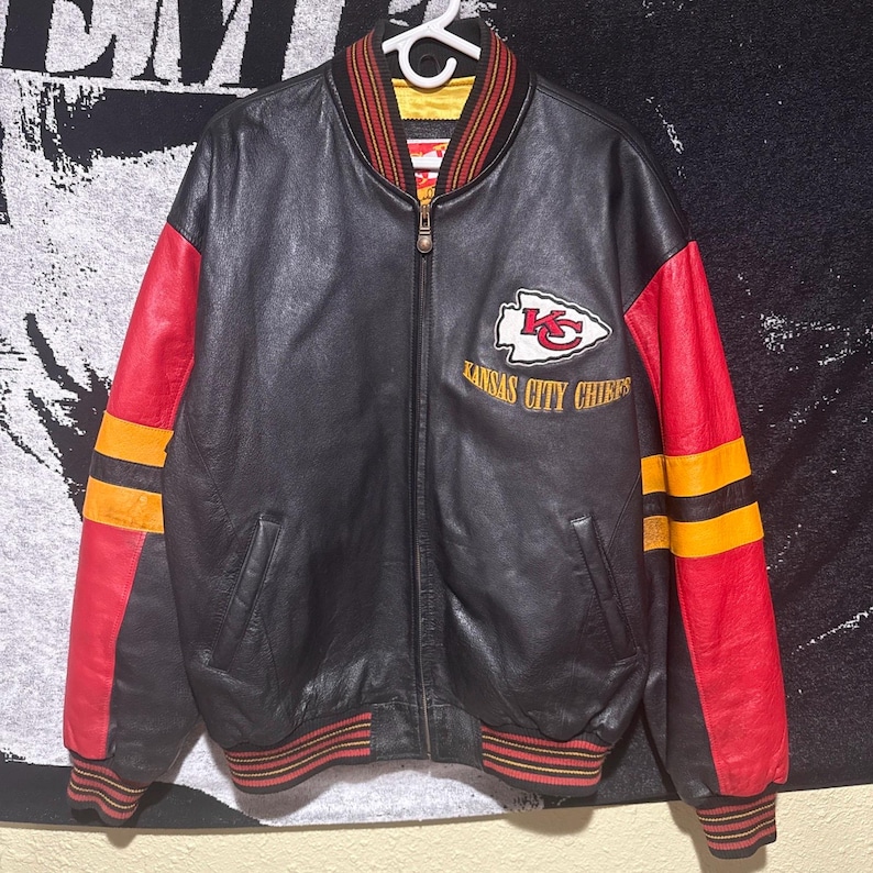 May include: Black leather bomber jacket with red and yellow accents. Features the Kansas City Chiefs logo and text on the chest. The jacket has a ribbed collar, cuffs, and hem with red and yellow stripes.