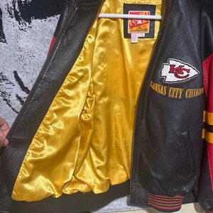 May include: Black and red leather jacket with a yellow satin lining. The jacket features the Kansas City Chiefs logo and lettering. The jacket has a ribbed collar, cuffs, and hem with red and yellow stripes. The jacket is hanging on a white hanger.
