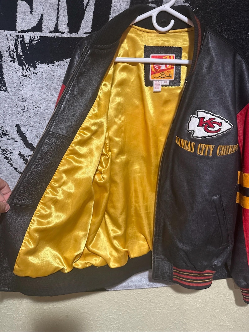 May include: Black and red leather jacket with a yellow satin lining. The jacket features the Kansas City Chiefs logo and lettering. The jacket has a ribbed collar, cuffs, and hem with red and yellow stripes. The jacket is hanging on a white hanger.