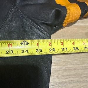 May include: Black leather jacket with yellow stripes on the upper arm. The jacket is measured with a yellow tape measure, showing a length of approximately 26 inches.