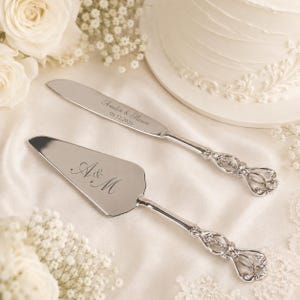 Puede incluir: Un juego de cuchillo y pala para pastel en plata con mangos ornamentados y calados. La hoja del cuchillo está grabada con el texto Amelia & Mason y la fecha 09.12.2026. La pala triangular presenta el monograma grabado A & M en letra cursiva.