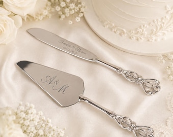Puede incluir: Un juego de cuchillo y pala para pastel en plata con mangos ornamentados y calados. La hoja del cuchillo está grabada con el texto Amelia & Mason y la fecha 09.12.2026. La pala triangular presenta el monograma grabado A & M en letra cursiva.