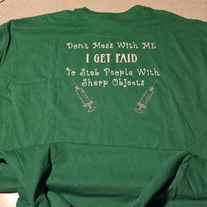 May include: Green t-shirt with white text that reads "Don't Mess With ME, I GET PAID, To Stab People With Sharp Objects." Two syringe graphics are below the text. The shirt is laid flat on a light-colored surface.
