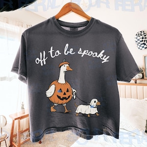 May include: A charcoal gray short-sleeve t-shirt featuring a white graphic of a goose wearing a pumpkin costume and a small dog wearing a ghost sheet. The text above the illustration reads off to be spooky in a white cursive script font.