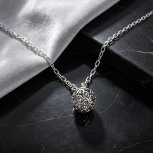 May include: A silver-toned necklace featuring a spherical pendant with an intricate, openwork filigree pattern. The pendant is suspended from a delicate, polished silver-toned chain with oval-shaped links, creating a cohesive metallic aesthetic.