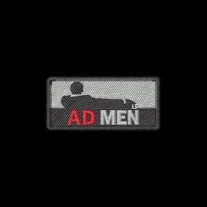 May include: A rectangular embroidered patch featuring a black silhouette of a person reclining on a couch against a light gray background. Below the figure, the text AD MEN is stitched in red and white thread with a black border surrounding it.