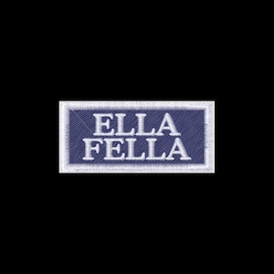 May include: A rectangular embroidered patch featuring a dark blue background with a white border. The text ELLA FELLA is stitched in white capital letters across the center of the patch. The embroidery has a visible diagonal thread texture.