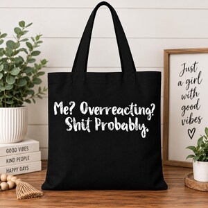 May include: A black tote bag featuring white text that reads, "Me? Overreacting? Shit Probably." The bag has two black shoulder straps and a rectangular shape. The lettering has a slightly distressed, textured appearance against the solid black fabric.