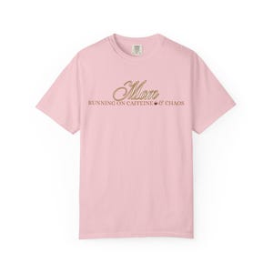 May include: A light pink t-shirt with the word "Mom" in a cursive gold font. Below the word "Mom" is the phrase "RUNNING ON CAFFEINE & CHAOS" in a smaller gold font. The shirt is a crew neck style.