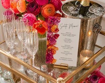 May include: A gold bar cart displays a floral arrangement of pink and orange flowers, champagne, wine glasses, and a menu. The menu reads "Salut Mama!" and lists wine and beverage options. A candle in a glass holder adds ambiance.