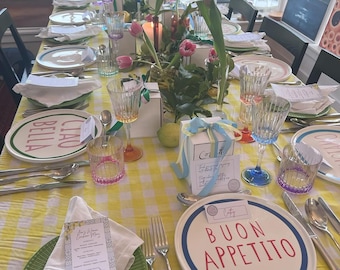 Puede incluir: Una mesa bellamente decorada con un mantel de cuadros amarillos y blancos, con platos con frases italianas, cristalería, cubertería y arreglos florales. El montaje de la mesa incluye servilletas blancas y cajas de regalo con cintas azules. Los tulipanes rosas y las velas añaden ambiente.